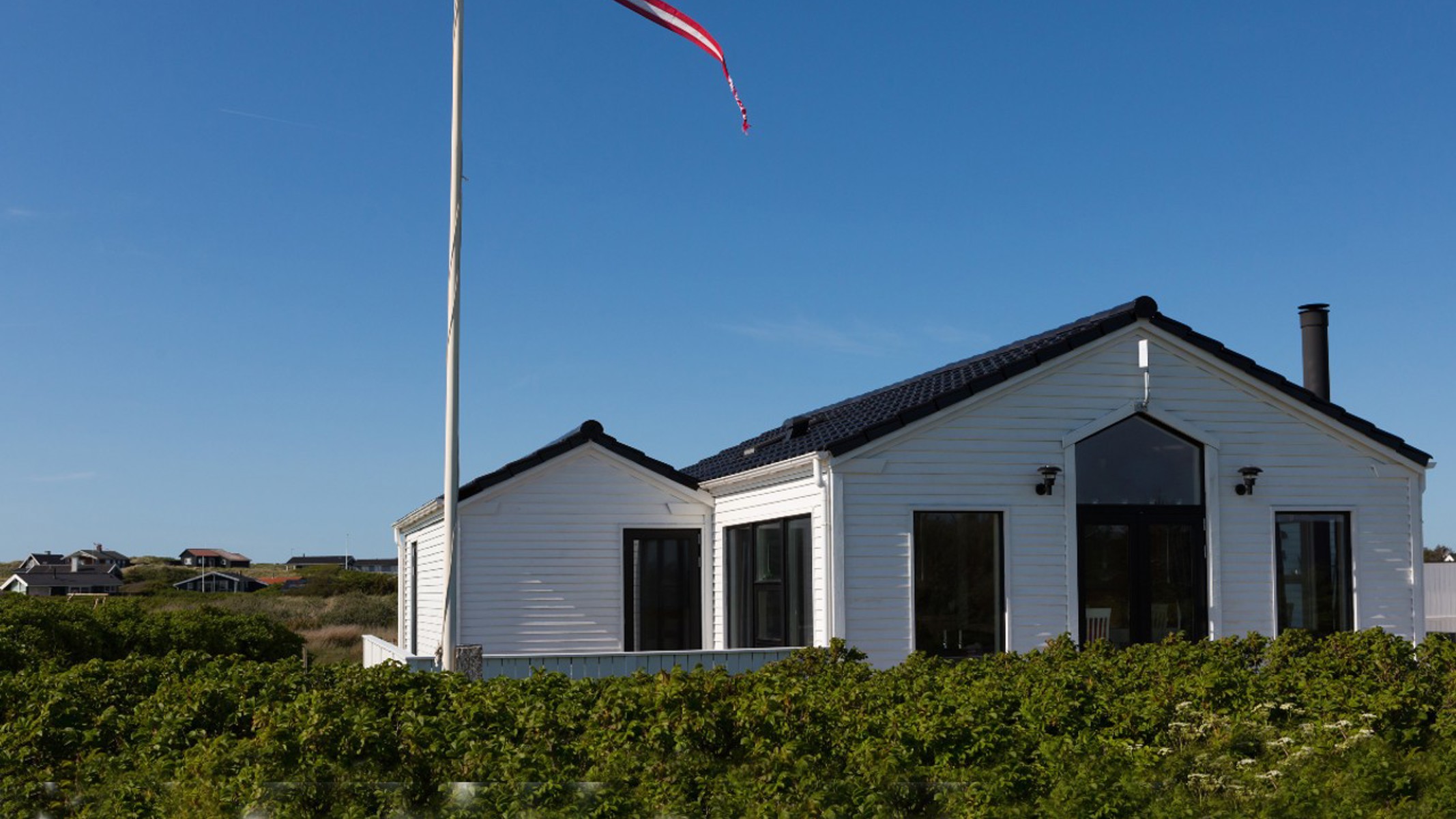 Hvidt sommerhus, beliggende i et sommerhusområde, med en flagstag med flag i.