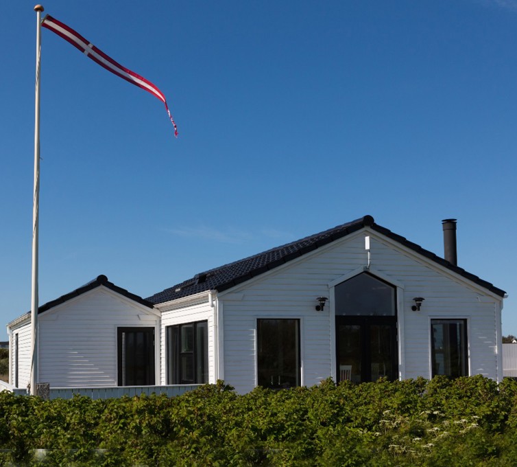 Hvidt sommerhus, beliggende i et sommerhusområde, med en flagstag med flag i.