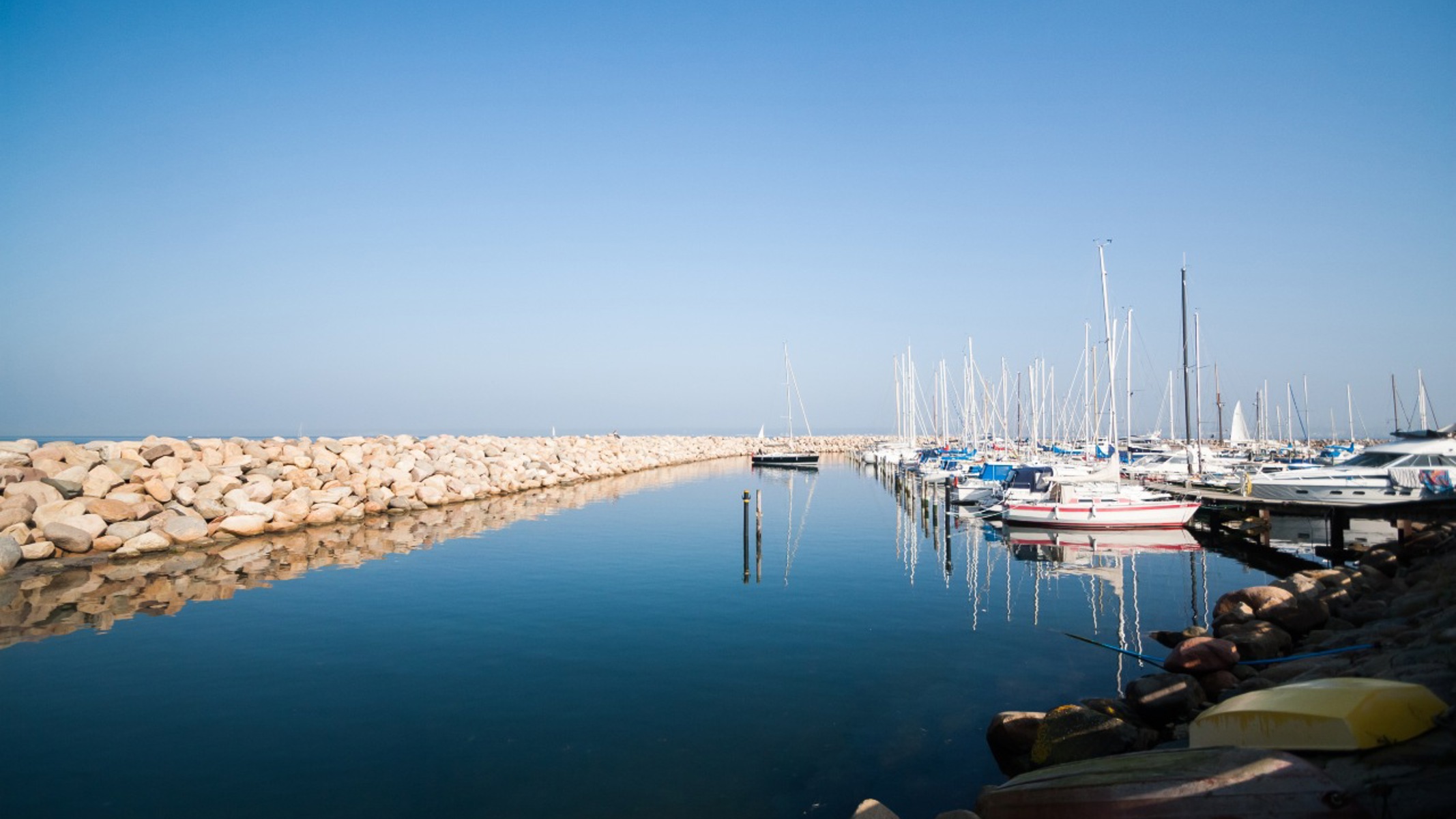 Lystbådehavn på en solskinsdag med spejlblankt vand, hvide sejlbåde fortøjet langs kajen og en stenmole, der beskytter havnen mod havet.