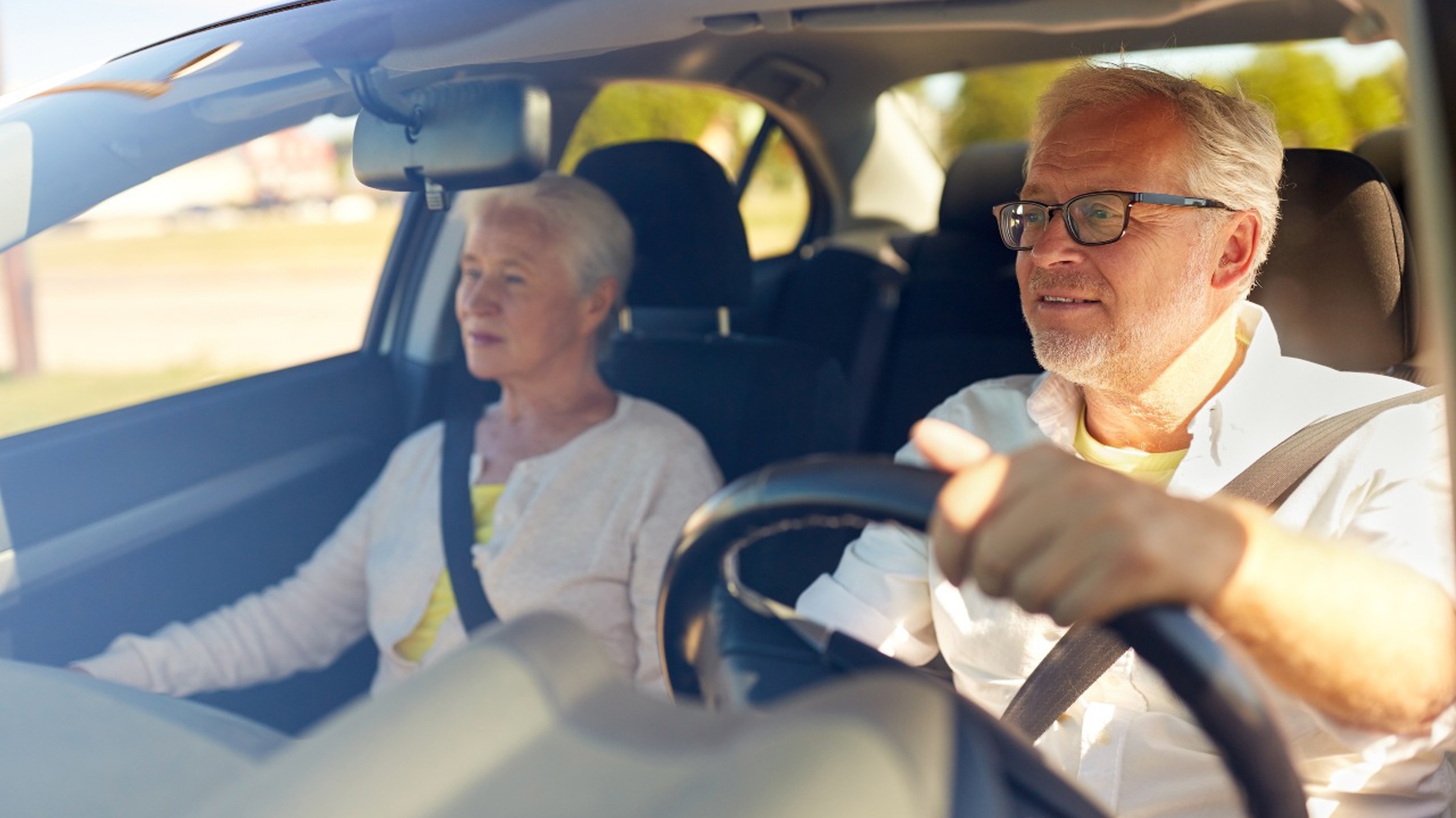 Ældre kvinde og ældre mand sidder i bil. Den ældre mand kører bilen. 