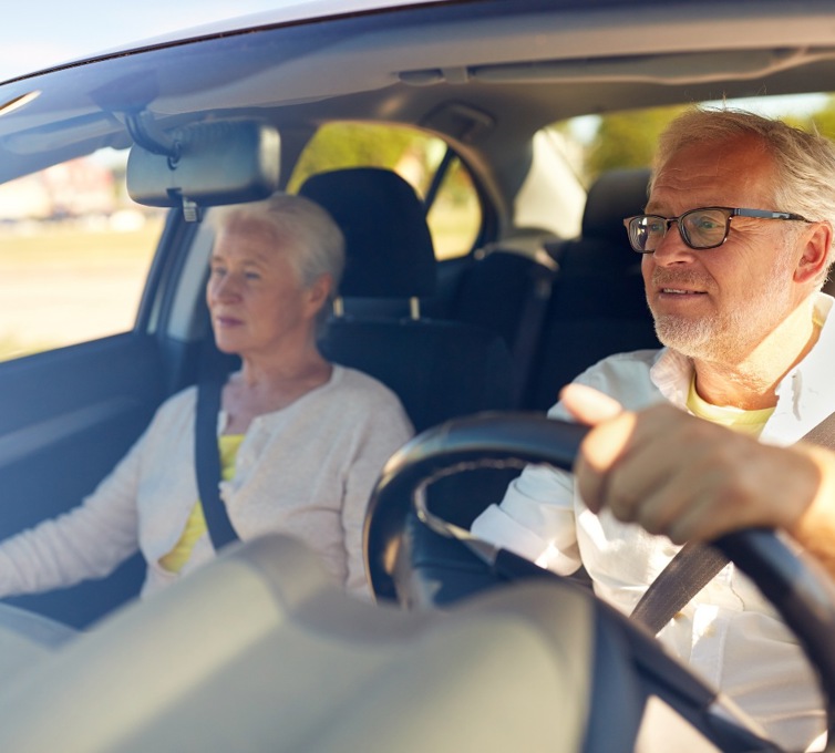 Ældre kvinde og ældre mand sidder i bil. Den ældre mand kører bilen. 