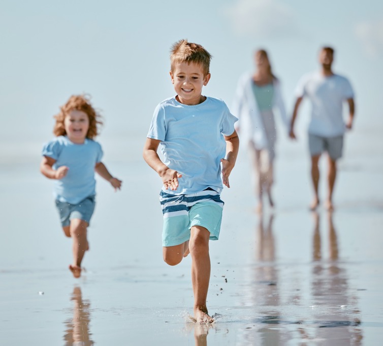 Lille dreng og lille pige løber glade på stranden, mens forældre går bagved og holder i hånd.