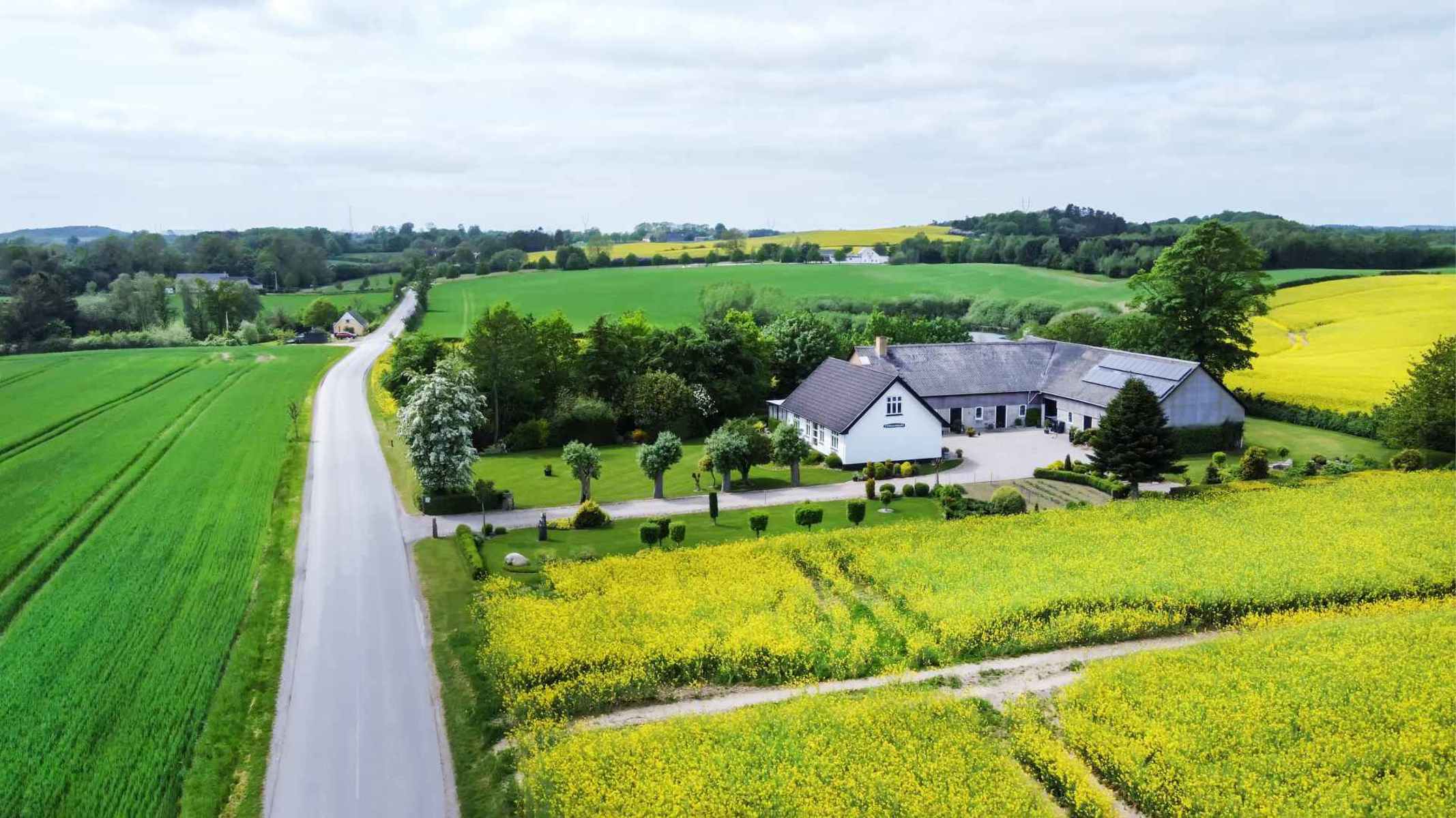 Hvid gård med omkringliggende marker og landevej.