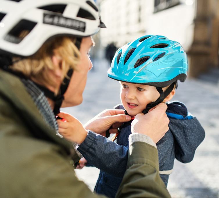 Kvinde med hvid cykelhjelm på, spænder en blå cykelhjelm på en lille dreng.