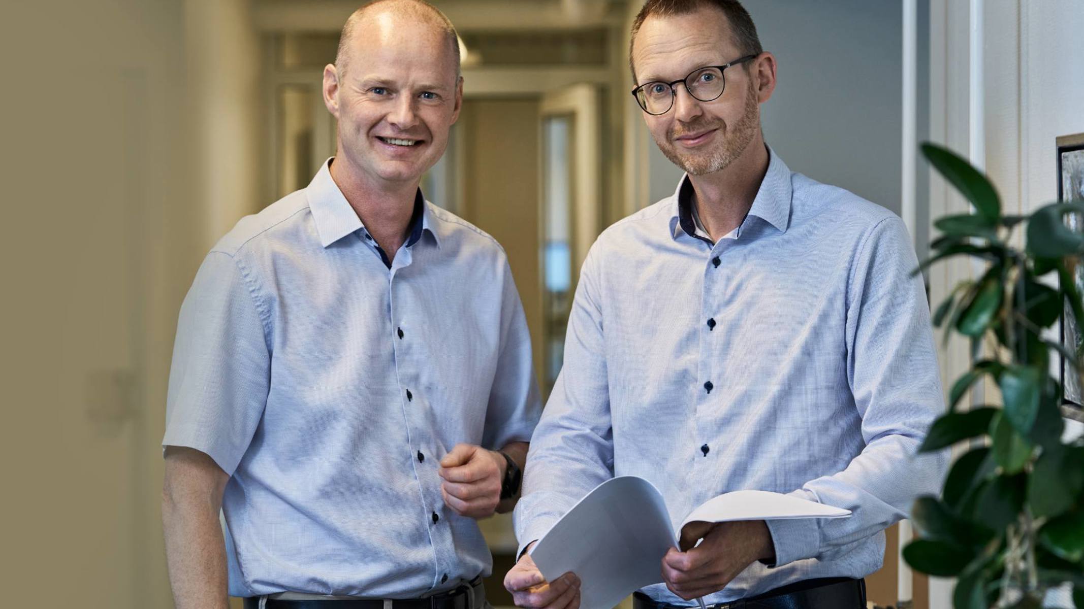 To forsikringsmæglere står ved siden af hinanden. Den ene står med papirer i hånden.