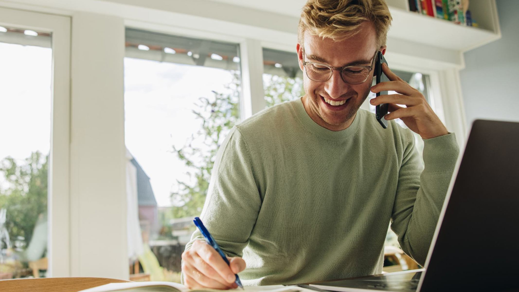 Ung mand sidder ved et bord med sin computer, mens han skriver ned i notesbog og taler i telefon.